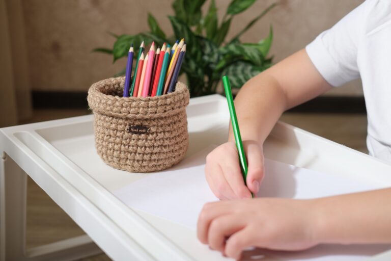 kid's hands drawing with color pencils and woven wicker basket for stationery storage.